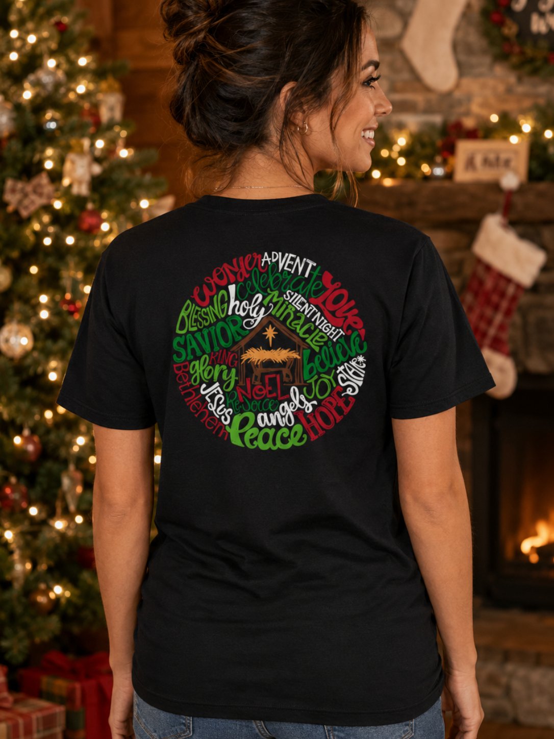 A woman wearing a black graphic t-shirt with a circular Christmas-themed text design on the back, standing in front of a decorated Christmas tree and a warm fireplace.
