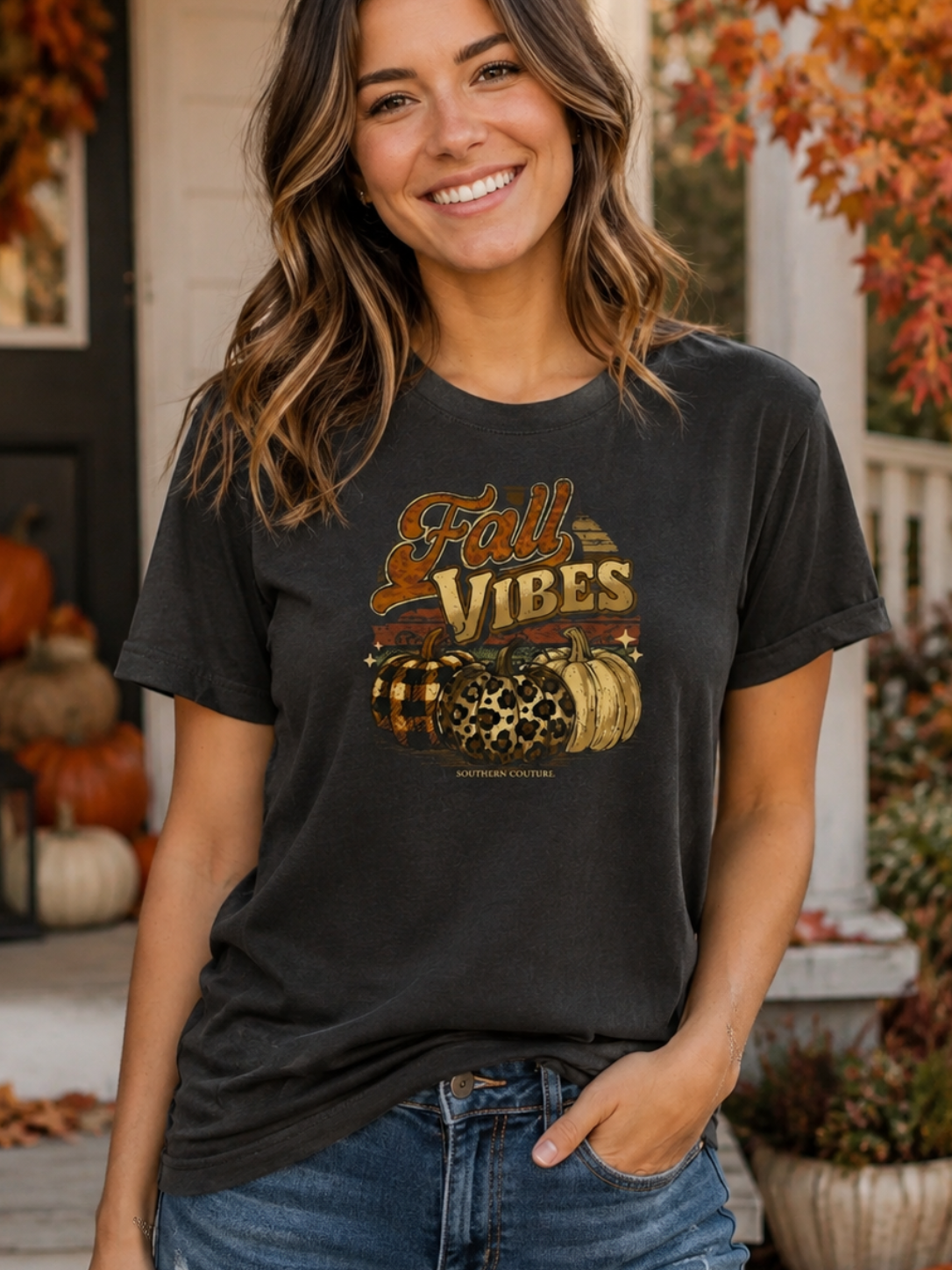 A woman smiling on a front porch wearing a dark charcoal gray graphic t-shirt with 'Fall Vibes' and pumpkins, paired with blue jeans.