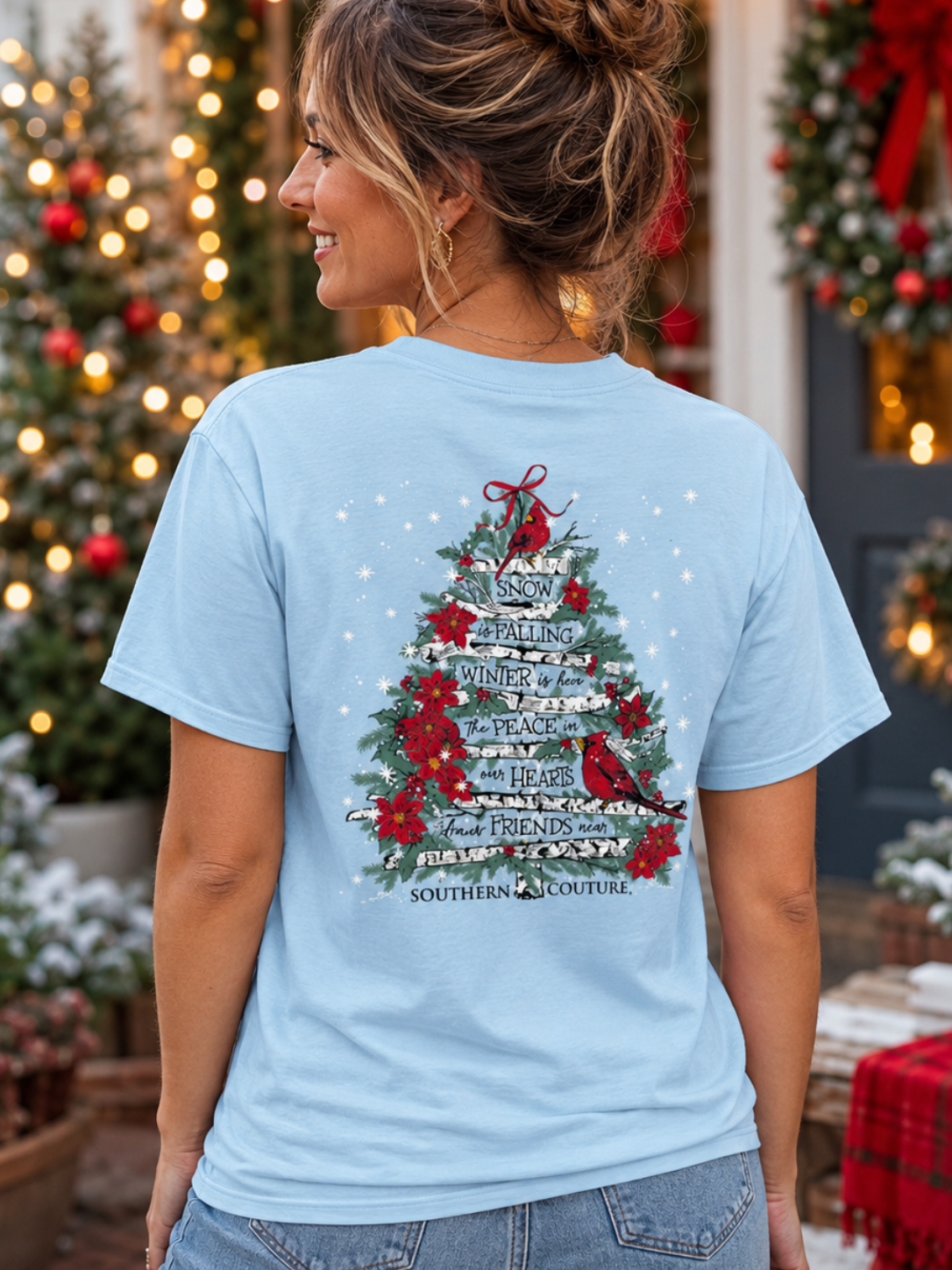 A woman wearing a light blue graphic t-shirt with a Christmas tree design, photographed from behind in a festive, decorated outdoor setting.