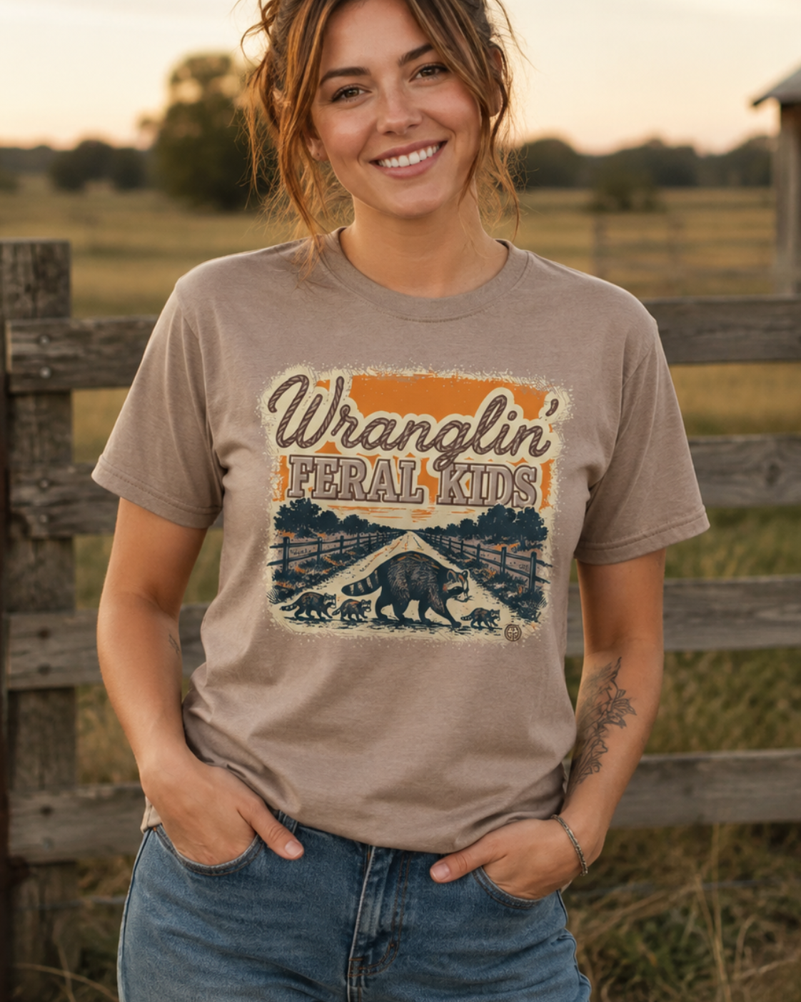 A woman wearing a beige graphic t-shirt with a 'Wrangler Feral Kids' bison design, standing in front of a rustic wooden fence at sunset.
