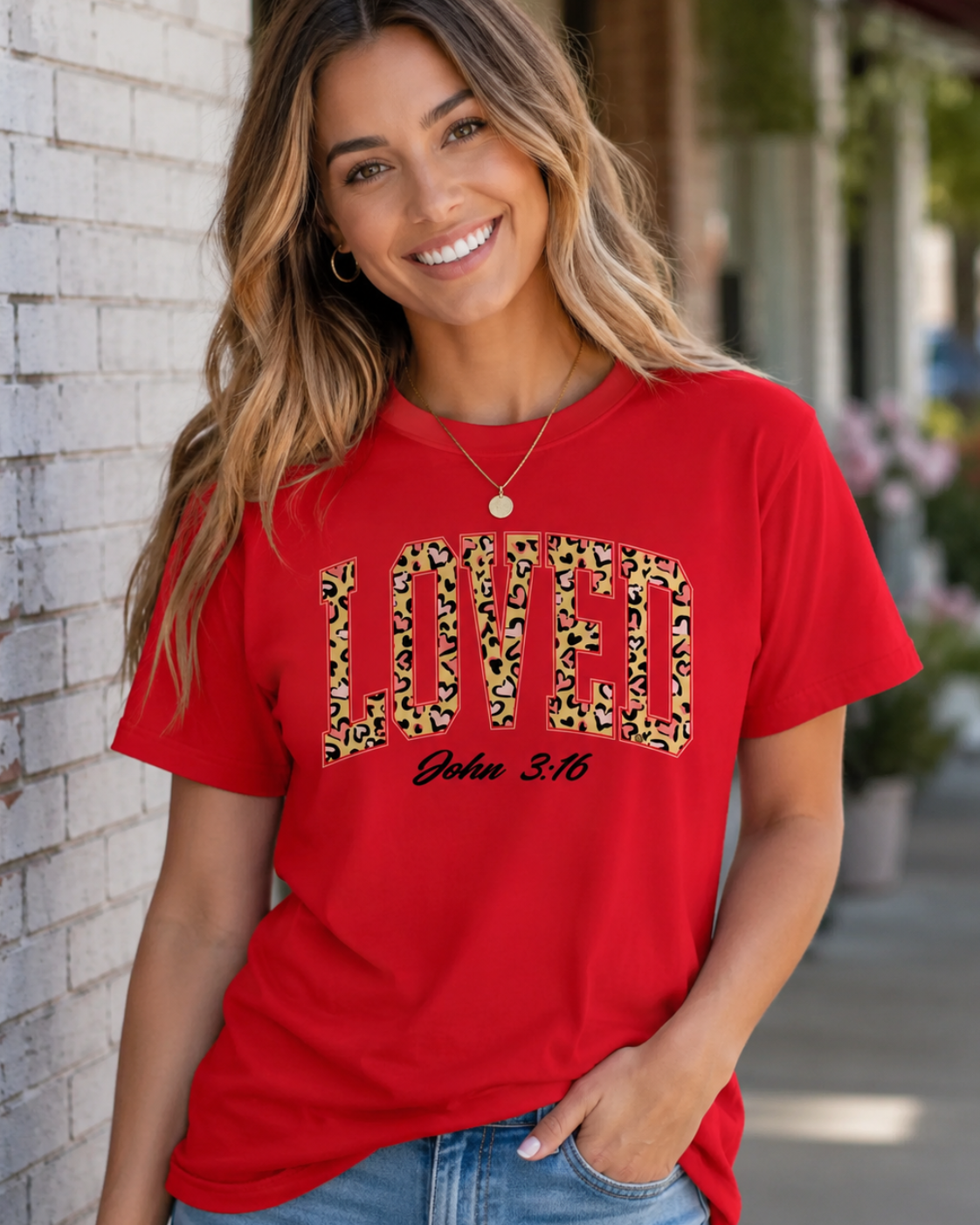 A woman wearing a red graphic t-shirt with 'LOVED' in leopard print and 'John 3:16' underneath, standing against a white brick wall.