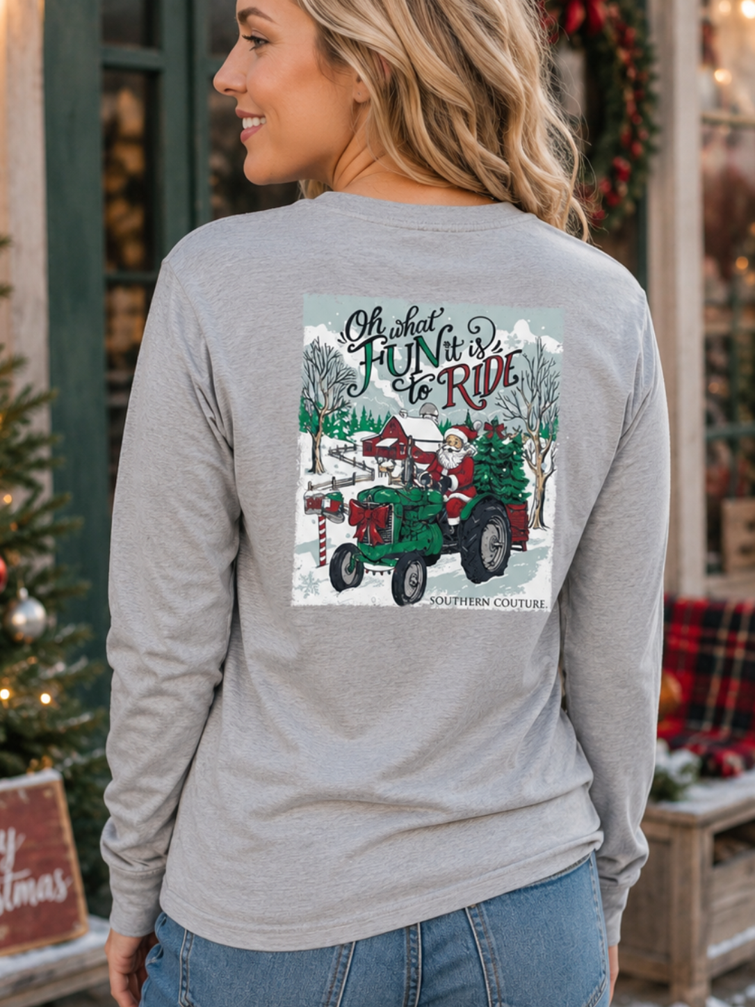 A woman wearing a light gray long-sleeve t-shirt with a large Christmas-themed graphic on the back, standing in front of a decorated holiday storefront.
