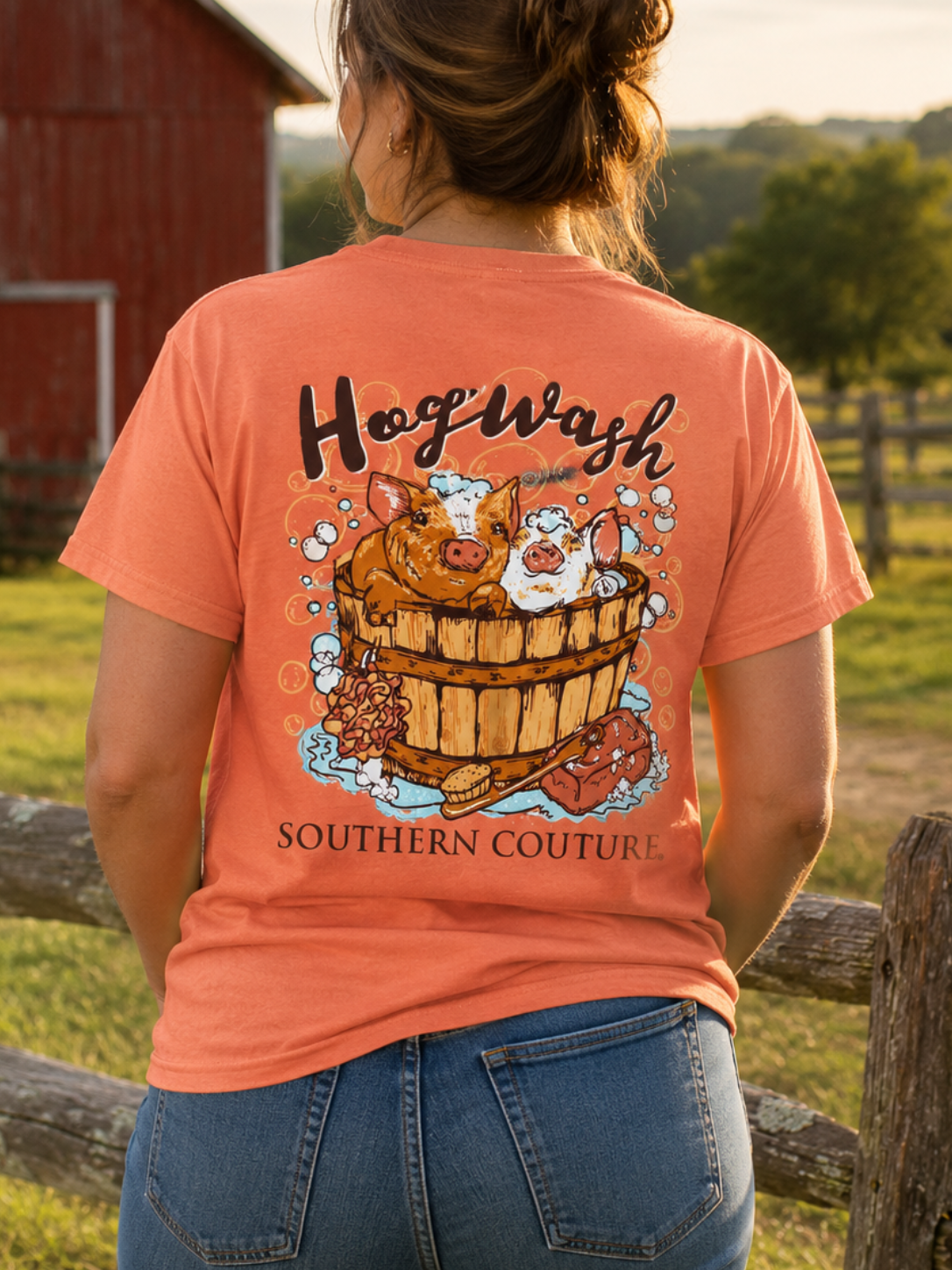 A woman stands outdoors in a rural setting with a red barn and wooden fence, viewed from behind. She is wearing a coral-orange graphic T-shirt with a large illustration of a cat in a wooden tub and the text 'Hogwash' and 'SOUTHERN COUTURE'.