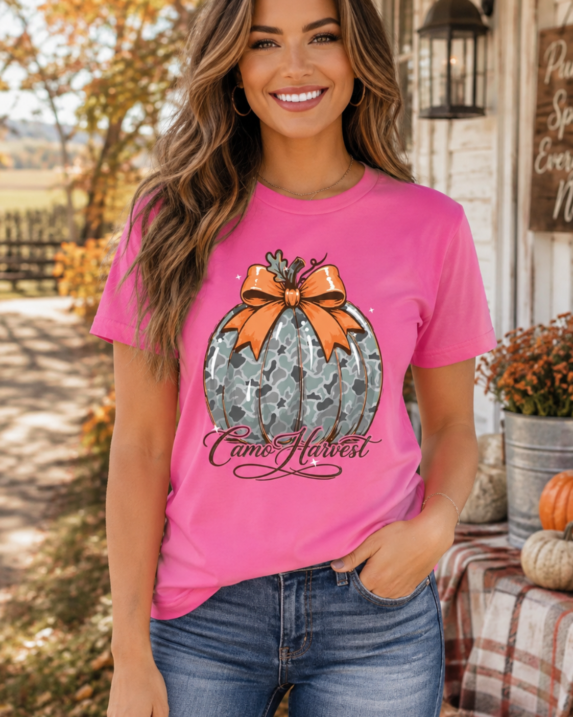 A woman wearing a bright pink graphic t-shirt with a fall harvest design, standing outdoors in front of a rustic house porch with pumpkins and autumn foliage.