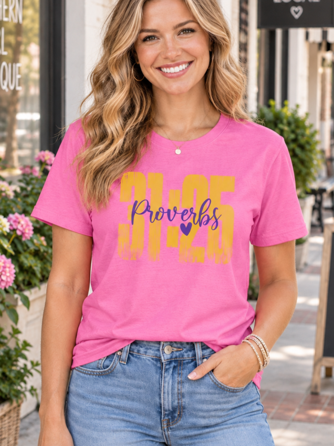 A smiling woman stands on a sidewalk wearing a bright pink graphic T-shirt with yellow and purple text reading 'Proverbs 31:25' and light blue jeans. Storefronts, flowers, and outdoor seating are visible in the background.