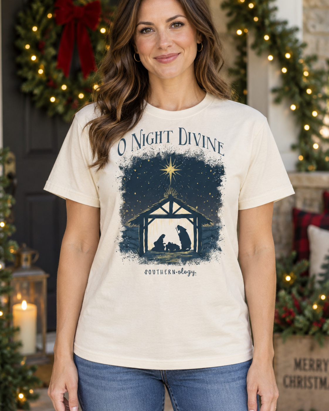 A woman wearing a cream-colored graphic t-shirt with a navy blue Christmas nativity design and the text 'O Night Divine' and 'EX FLORESCENDO'. She is standing in front of a decorated holiday doorway with wreaths, lights, and candles.
