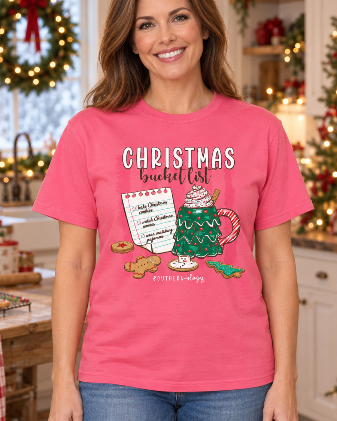 A smiling woman stands in a decorated holiday kitchen wearing a bright pink graphic T-shirt with the text 'CHRISTMAS bucket list' and a festive mug, cookie, and candy cane illustration. She is also wearing blue jeans.