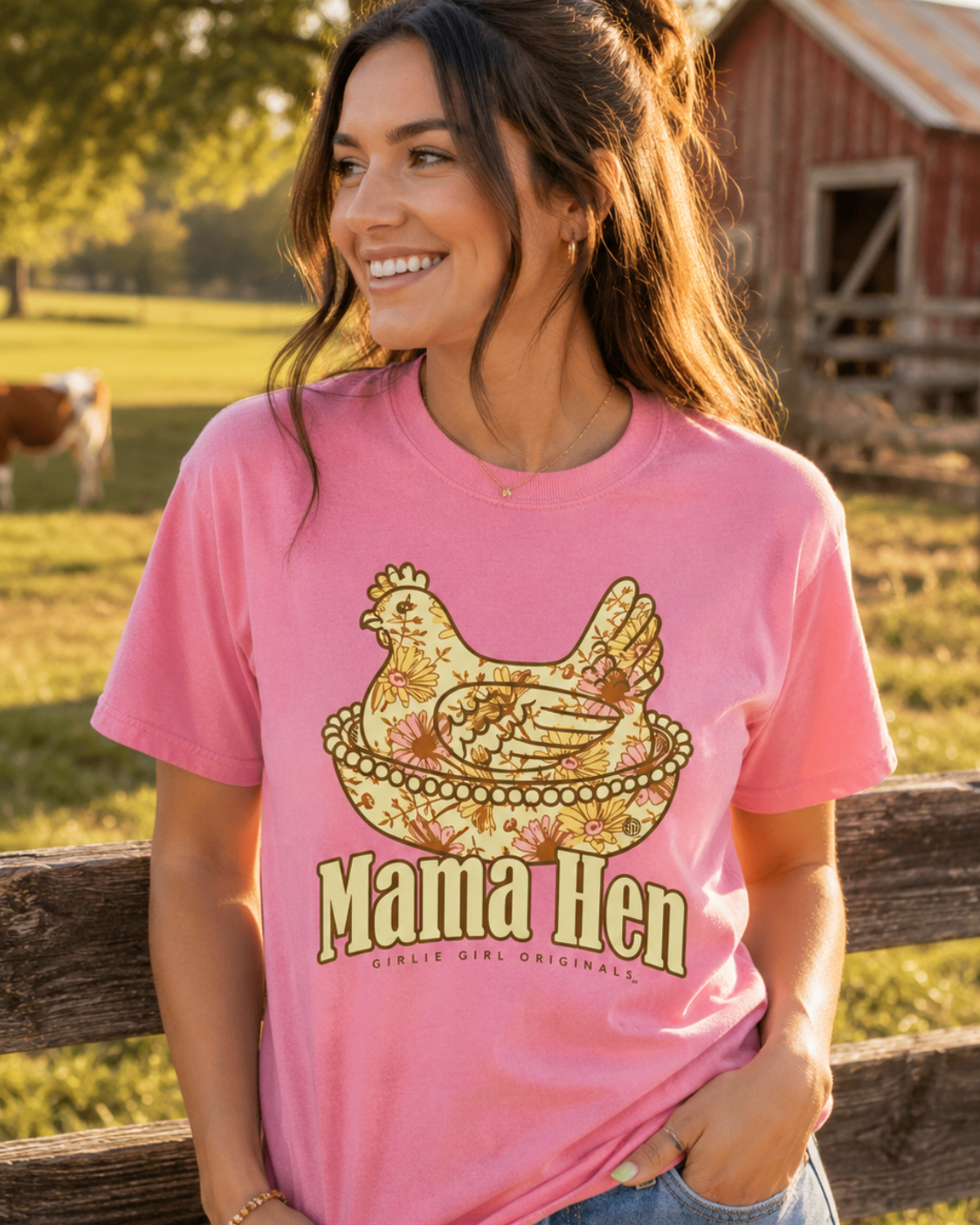 A woman wearing a pink graphic t-shirt with a 'Mama Hen' design, standing outdoors in front of a wooden fence with a red barn and cow in the background.