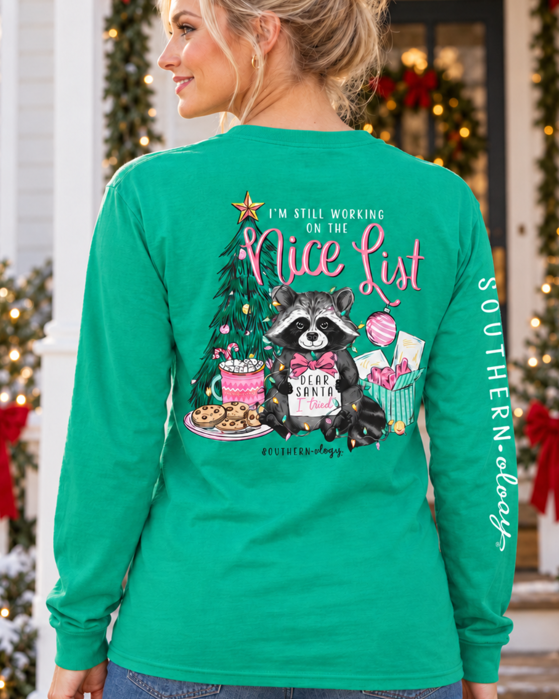 A woman wearing a bright green long-sleeve shirt with a large Christmas-themed graphic on the back, standing in front of a decorated holiday porch. The shirt features a raccoon holding a Santa hat next to a Christmas tree with the text 'I'm Still Working On My Nice List' and 'SOUTHERN Comfort' on the sleeve.
