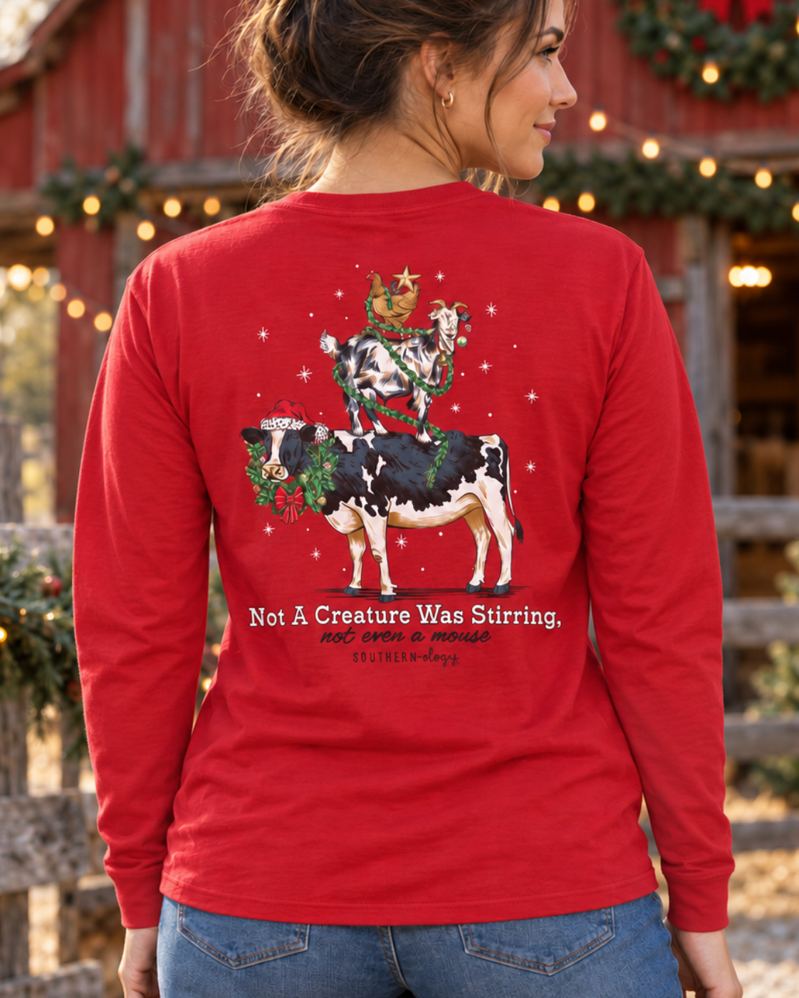 A woman shown from behind wearing a bright red long-sleeve t-shirt with a festive Christmas graphic of a cow riding a horse with a wreath, set against a blurred background of a decorated red barn and wooden fence.