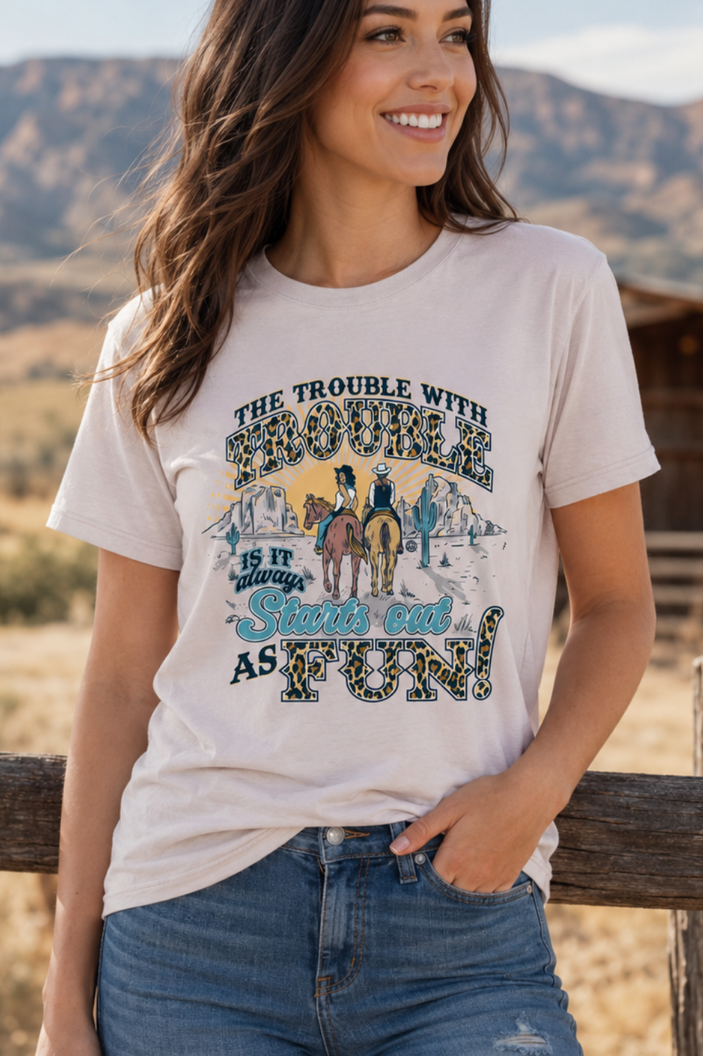 A woman wearing a light beige graphic t-shirt and blue jeans, leaning against a rustic wooden fence in a sunny, dry outdoor landscape with mountains and a windmill in the background.