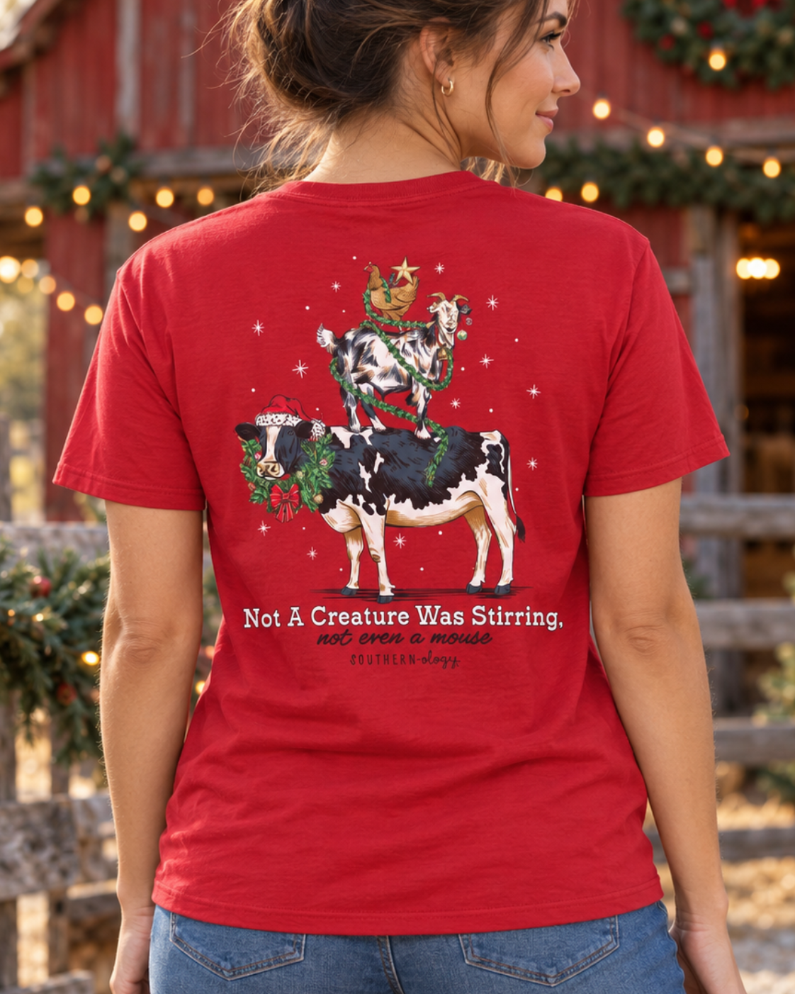 A woman stands outdoors in front of a decorated red barn wearing a red graphic T-shirt. The back of the shirt shows a Christmas-themed cow illustration with the text 'Not A Creature Was Stirring.