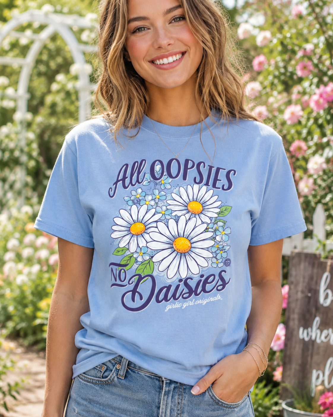 A woman wearing a light blue graphic t-shirt with a daisy design and the text 'All OOPSIES No Daisies' in a garden setting.
