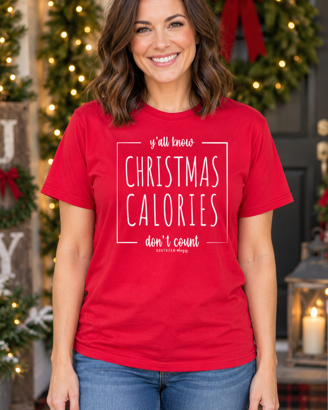 A woman wearing a bright red graphic t-shirt with white text that reads 'y'all know CHRISTMAS CALORIES don't count' by The Joy Shop, posed in front of a festive Christmas decorated doorway.