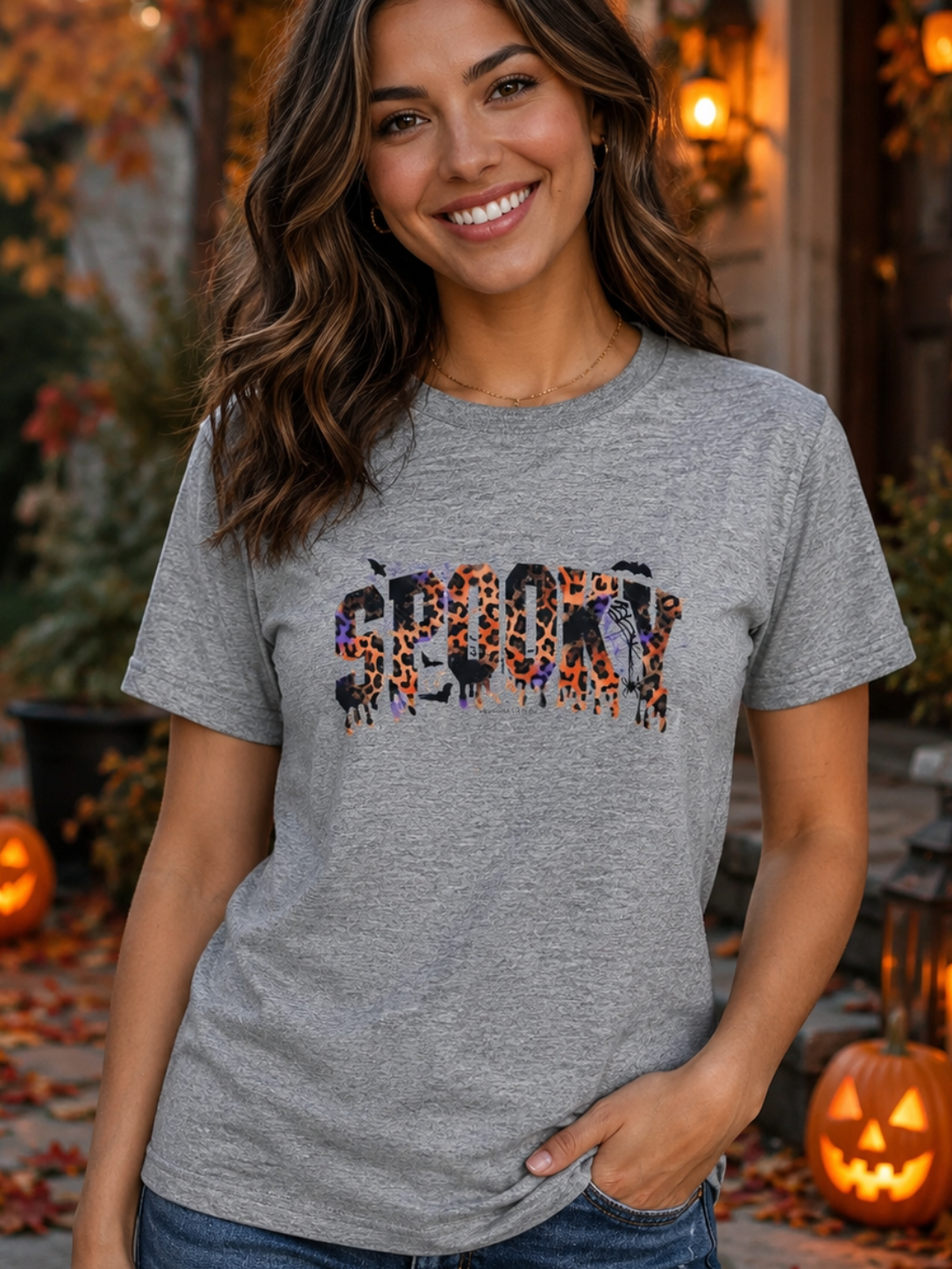 A woman wearing a heather gray graphic t-shirt with a 'SCARY' text design filled with Halloween motifs, posed outdoors in a cozy, warmly lit autumn setting with pumpkins and string lights.