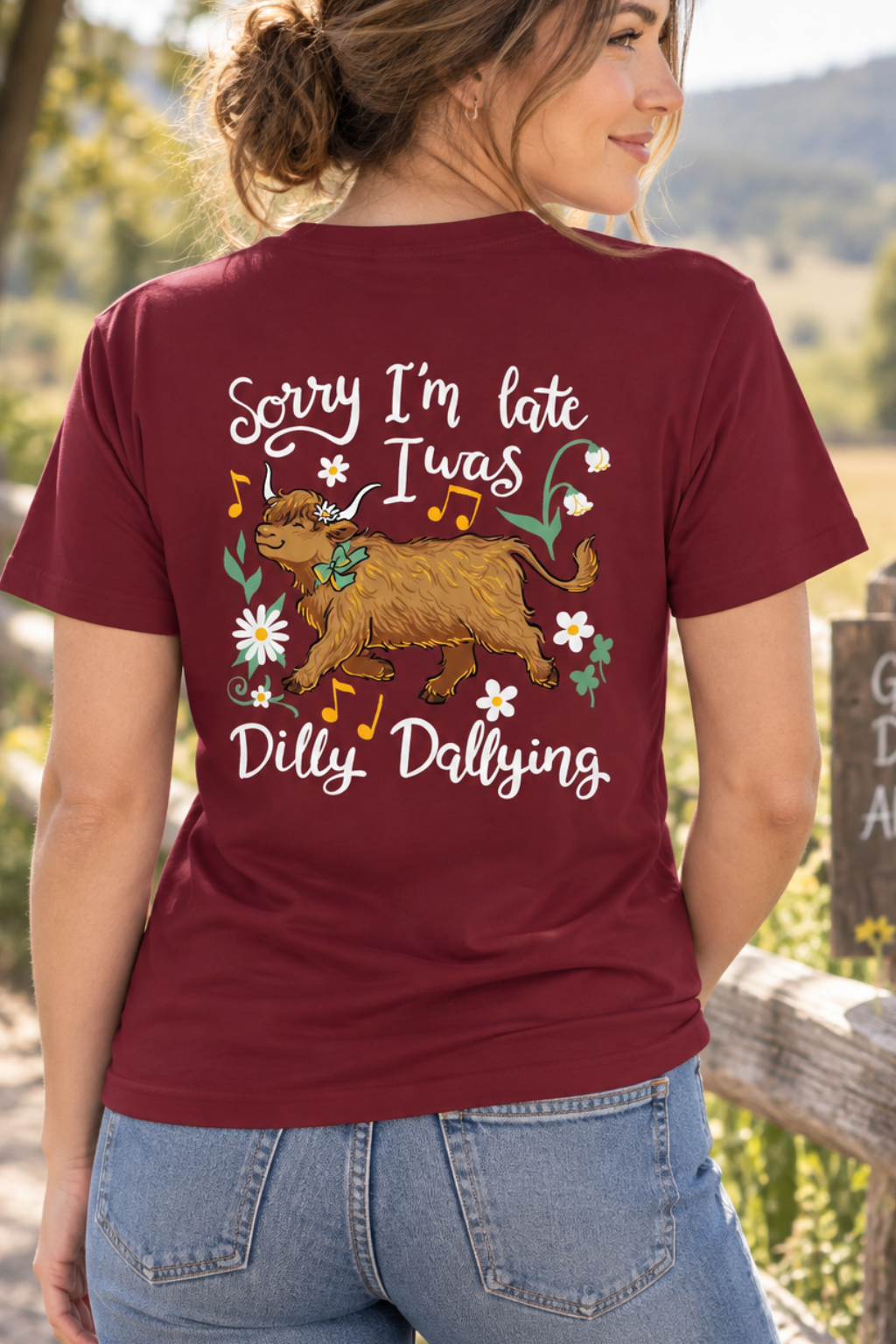 A woman wearing a maroon t-shirt with a humorous graphic on the back, standing outdoors next to a wooden fence with a sign that reads 'Good Days Ahead'. The graphic features a brown dog, white flowers, and white text reading 'Sorry I'm late Iwas Dilly Dallying'.
