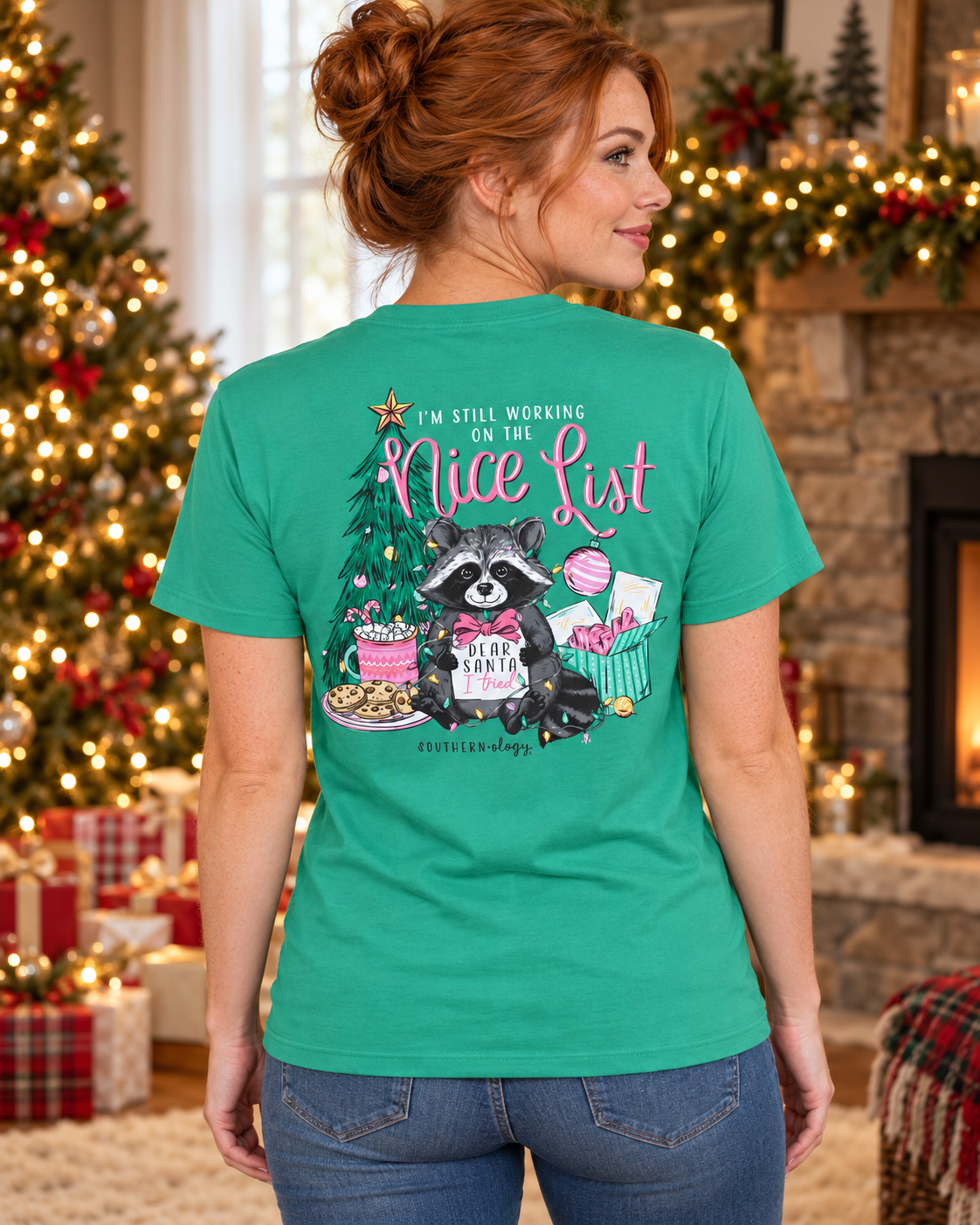 A woman wearing a green graphic t-shirt, viewed from behind, standing in a cozy, decorated Christmas living room with a fireplace and tree in the background. The shirt features a cartoon raccoon sitting on a Christmas tree with the text 'I'M STILL WORKING ON THE Nice List'.