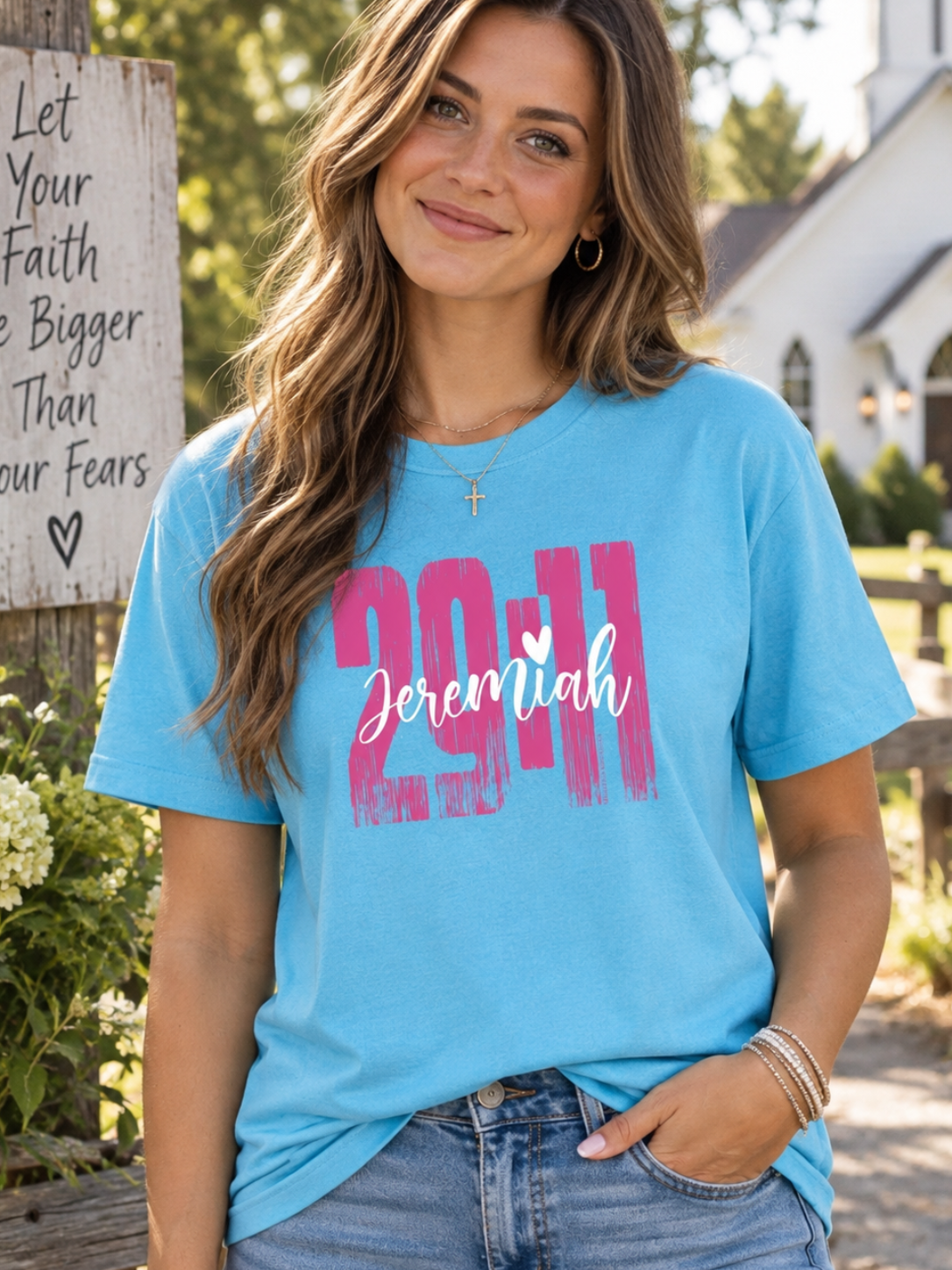 A woman wearing a light blue graphic t-shirt with '29:11 Jeremiah' printed on the front, standing outdoors in front of a wooden sign and a white church.