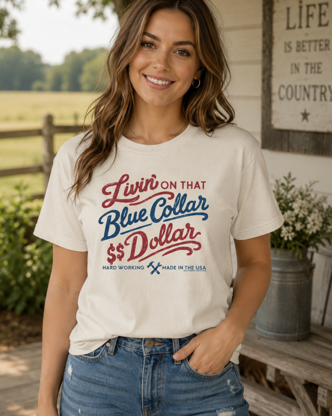 A woman wearing a cream-colored graphic t-shirt with red and blue text that reads 'Livin' ON THAT Blue Collar $$ Dollar' and 'MAD WORKING MADE IN THE USA', paired with blue jeans, standing in front of a rustic country-style house porch.