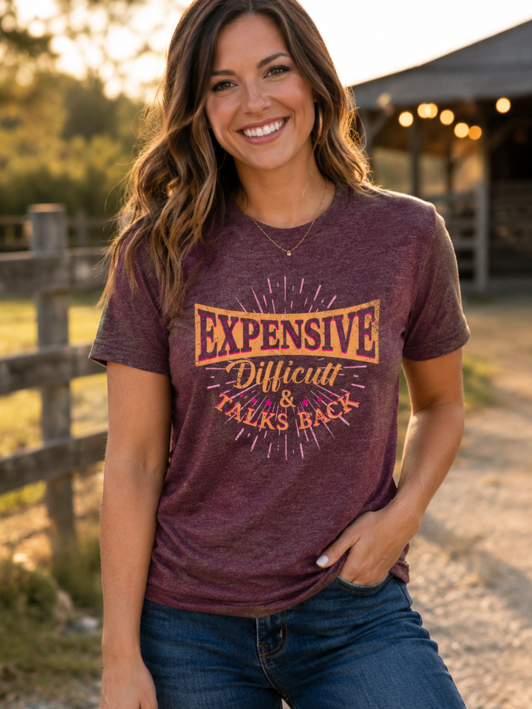A woman wearing a heathered maroon graphic t-shirt with the text 'EXPENSIVE Difficult &amp; TALKS BACK' in orange and yellow, paired with blue jeans, standing outdoors in front of a rustic barn at sunset.