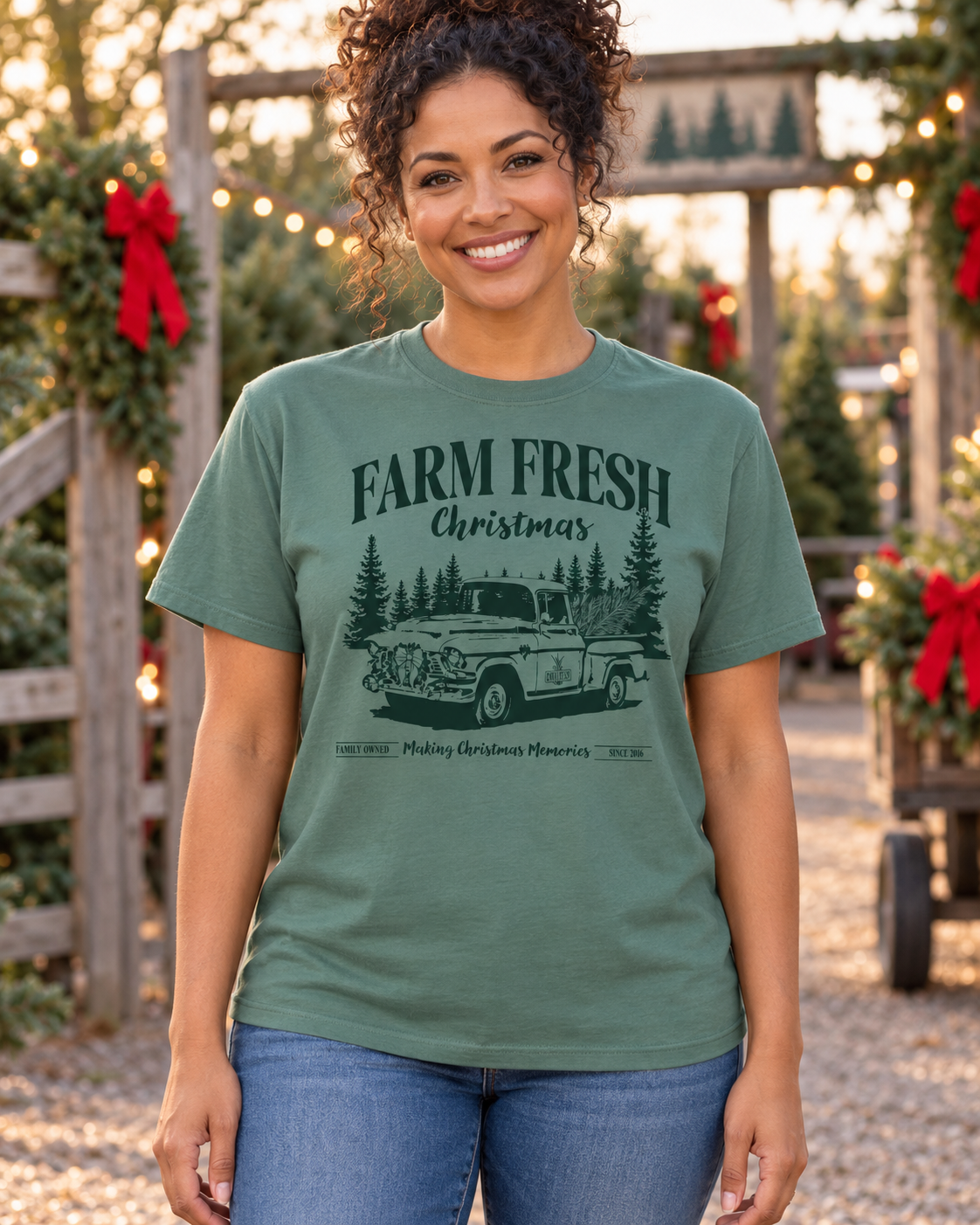 A woman wearing a sage green graphic t-shirt with a vintage truck and pine tree design that reads 'FARM FRESH Christmas'. The background shows a festive outdoor holiday scene with Christmas trees and warm string lights.