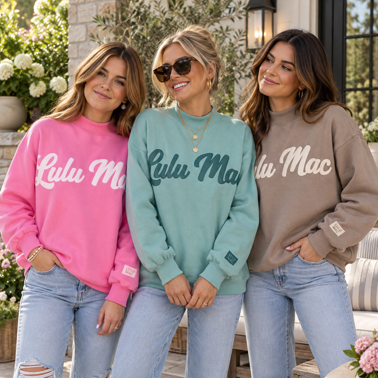 Three women standing together outdoors wearing oversized crewneck sweatshirts with 'Lulu Mac' written across the front in contrasting colors: pink, mint green, and taupe. They are paired with light-wash jeans.