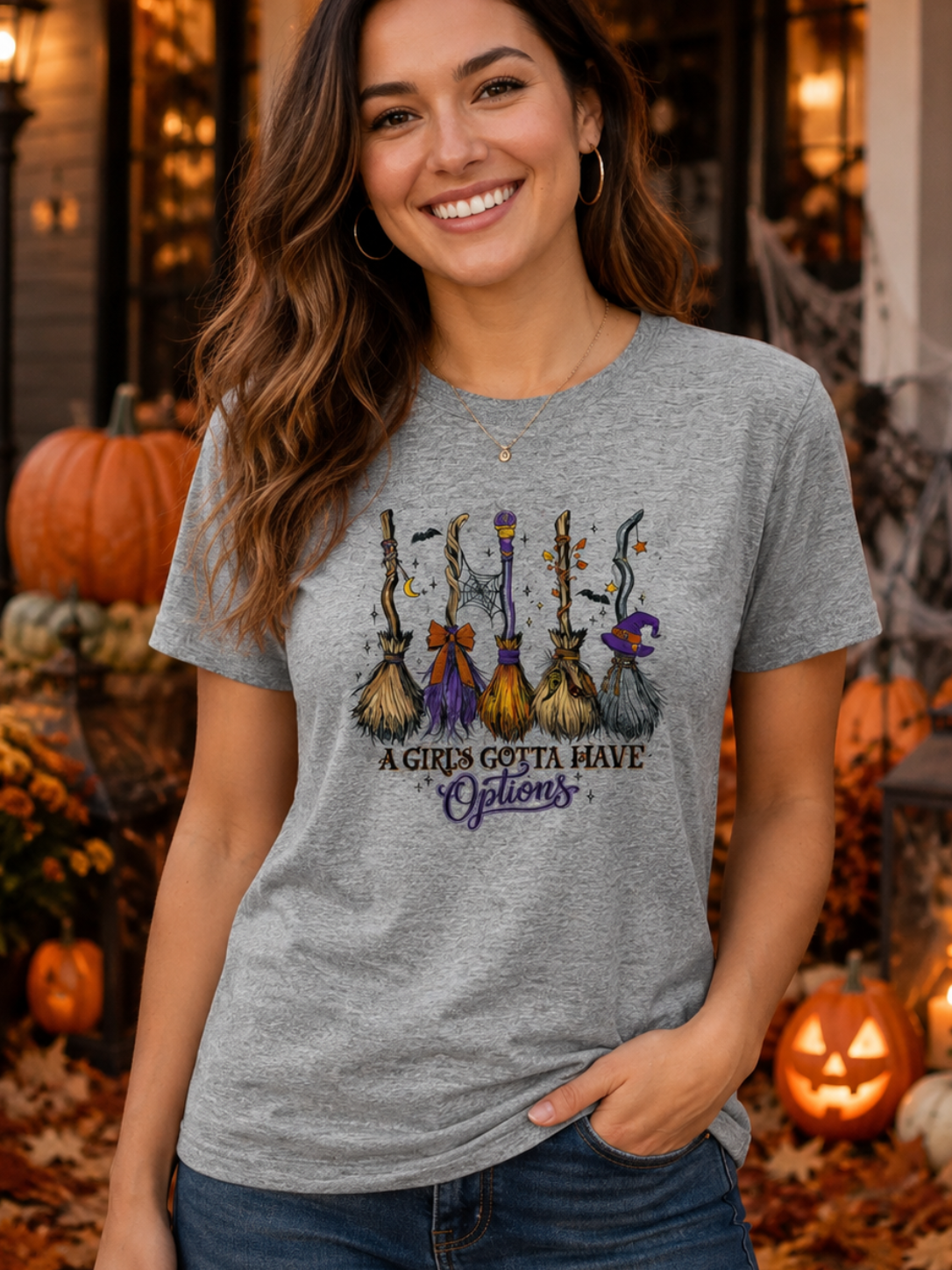 A woman wearing a heather gray graphic t-shirt with a Halloween-themed design of three witches on broomsticks and the text 'A GIRL'S GOTTA HAVE Options', posed outdoors in a decorated autumn porch setting with pumpkins and fall leaves.