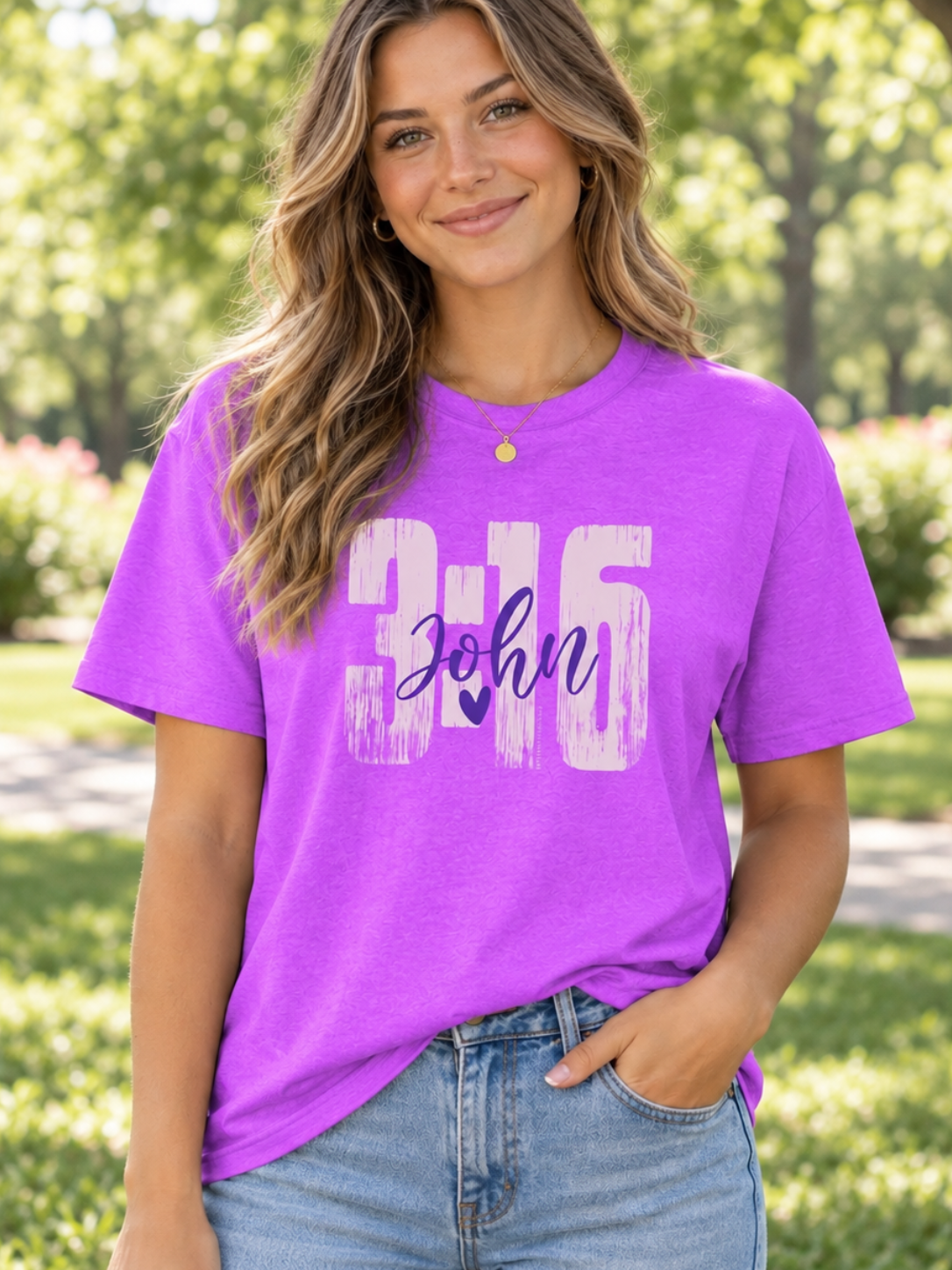 A woman wearing a bright purple graphic t-shirt with '316 John' printed on the front, paired with light blue jeans, standing outdoors in a sunny park with green trees in the background.