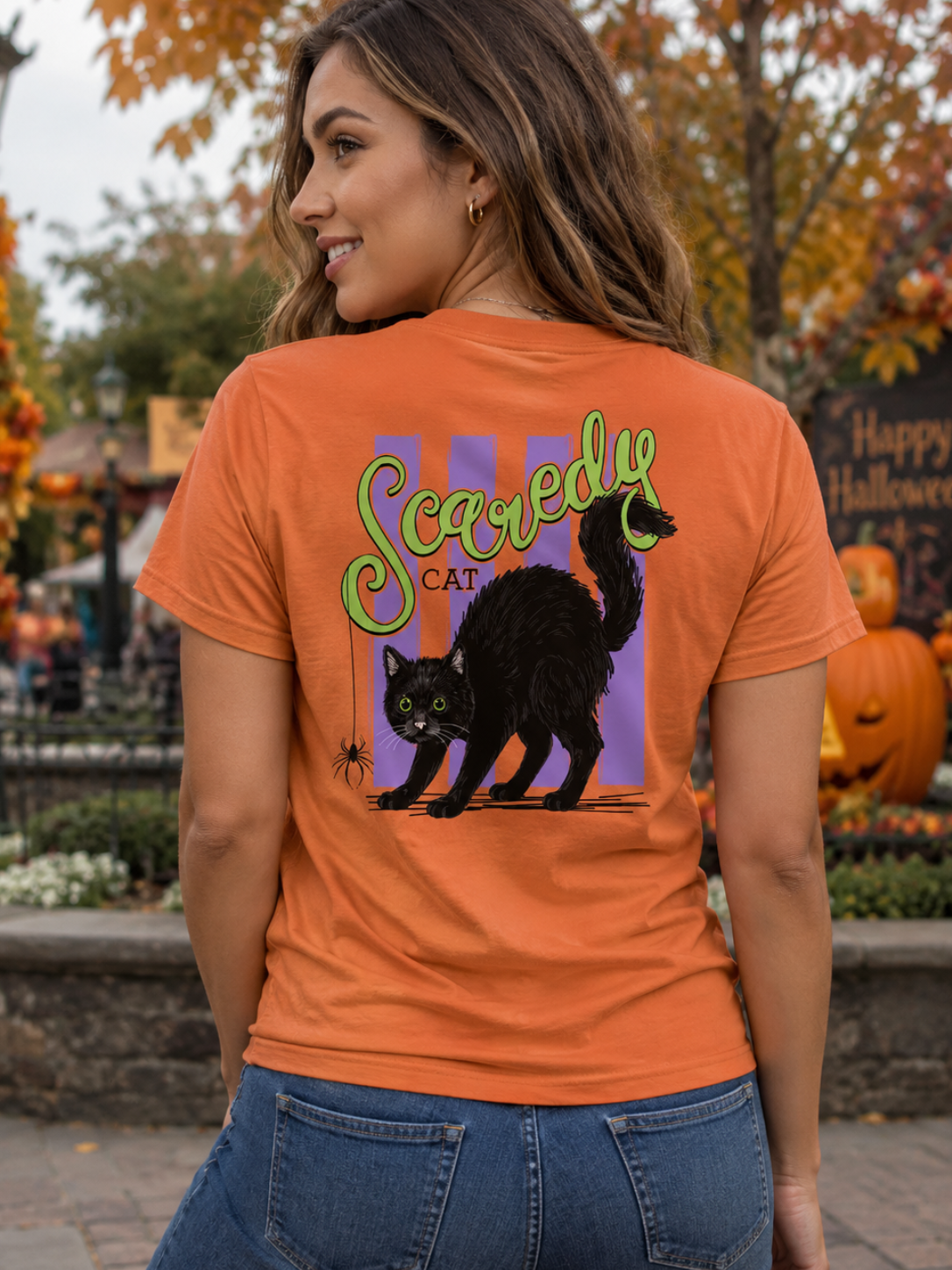 A woman wearing an orange graphic t-shirt with a black cat and 'Scaredy Cat' design, photographed from behind in an outdoor Halloween-themed setting with pumpkins and autumn foliage.