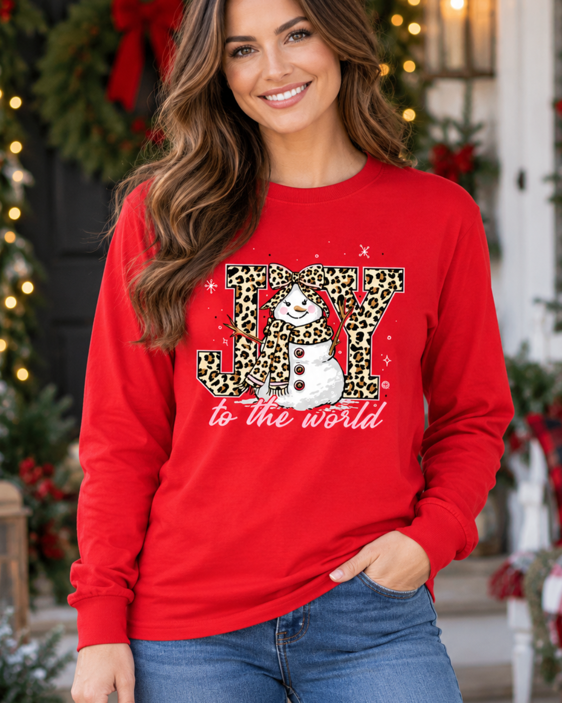 A woman wearing a bright red long-sleeve crewneck sweatshirt with a festive Christmas graphic that reads 'JOY to the world' in leopard-print and white script, paired with blue jeans, posed in front of a decorated holiday porch.