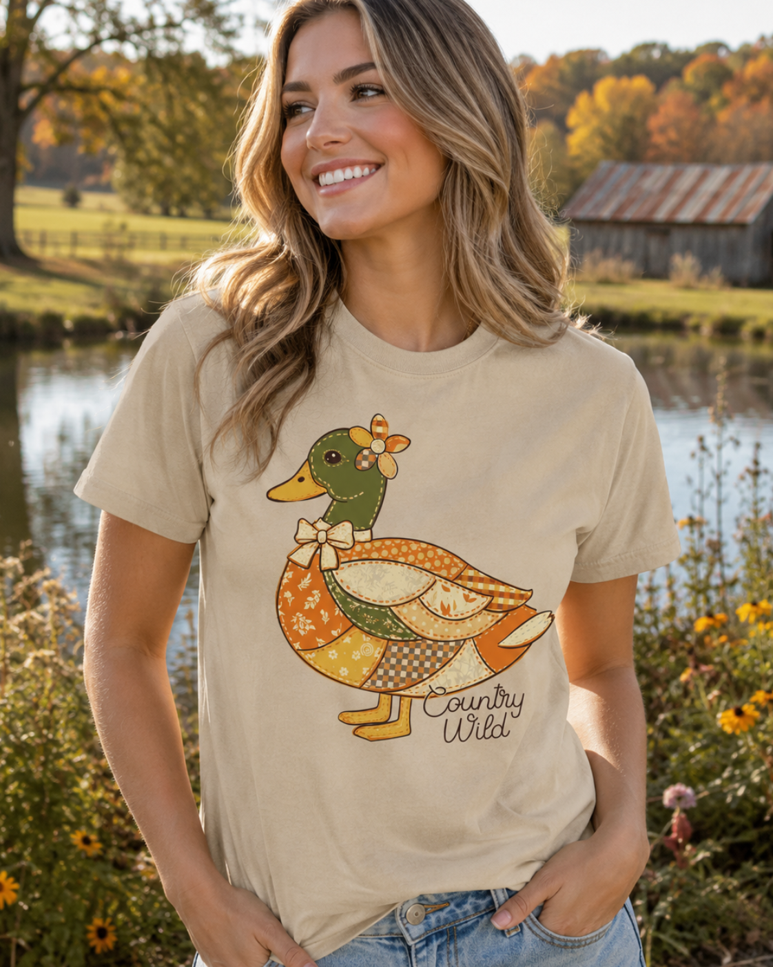 A woman wearing a beige graphic t-shirt with a colorful 'Country Wild' duck design, standing outdoors near a pond with autumn trees and a rustic barn in the background.