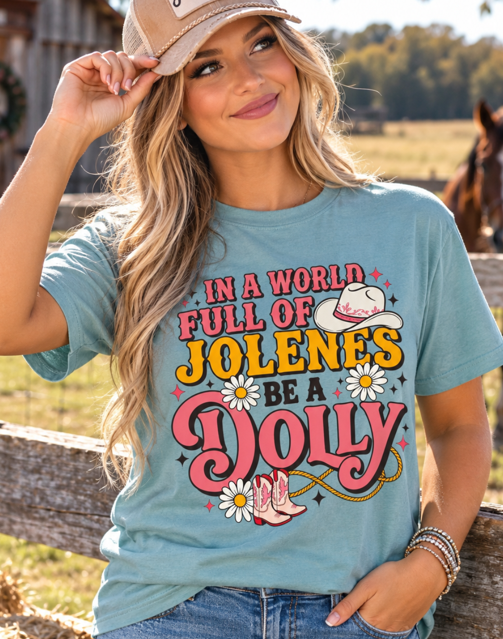 A woman wearing a light blue graphic t-shirt with a western-themed print that reads 'IN A WORLD FULL OF JOLENES BE A DOLLY' with cowboy boots, daisies, and a hat, paired with a tan 'Howdy' baseball cap and blue jeans in a rustic barn setting.