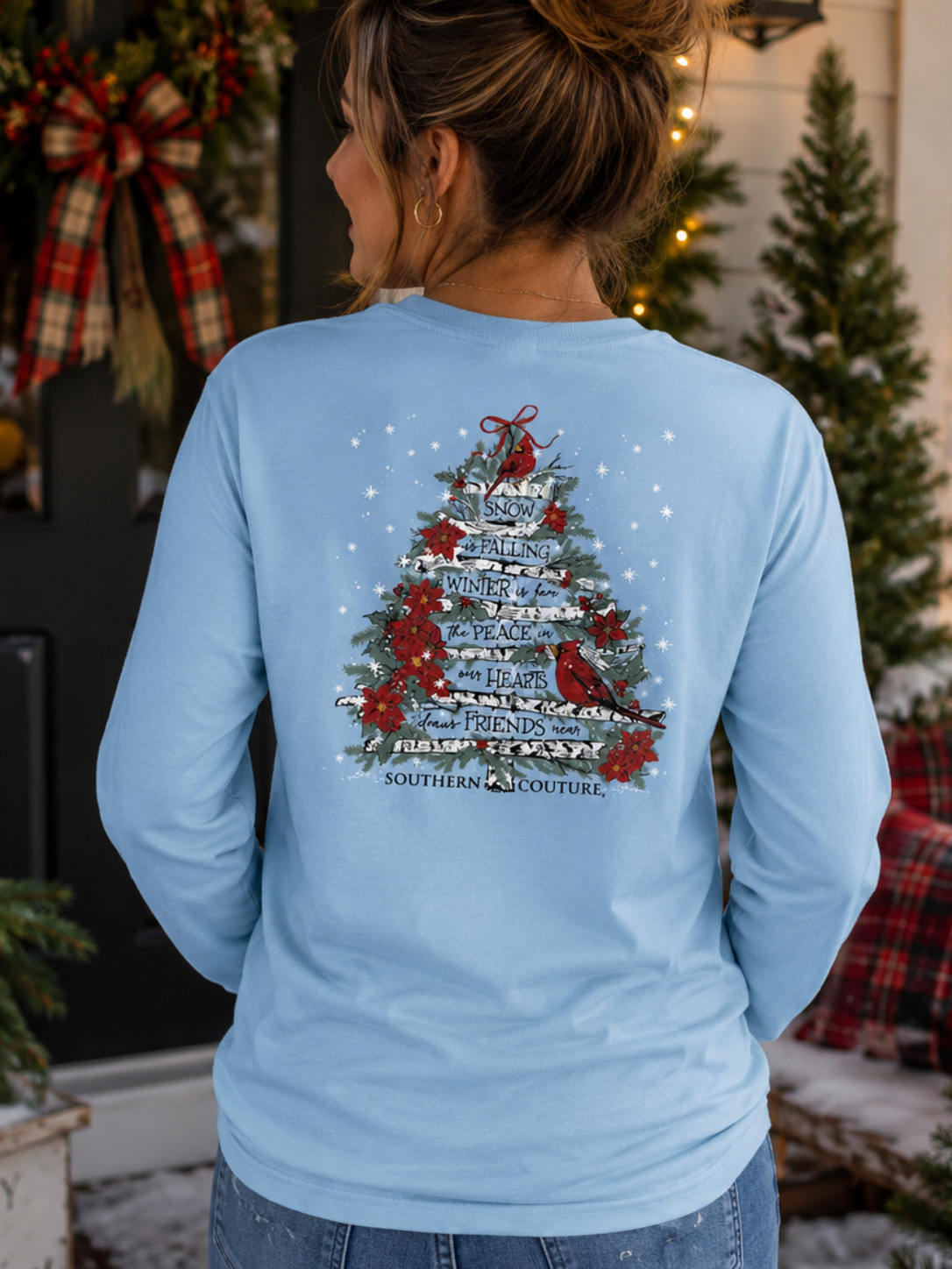 A woman viewed from behind wearing a light blue long-sleeve t-shirt with a large Christmas tree graphic on the back. The graphic features the words 'NORTH CHRISTMAS' and 'MERRY CHRISTMAS' in white, decorated with red birds and ornaments.