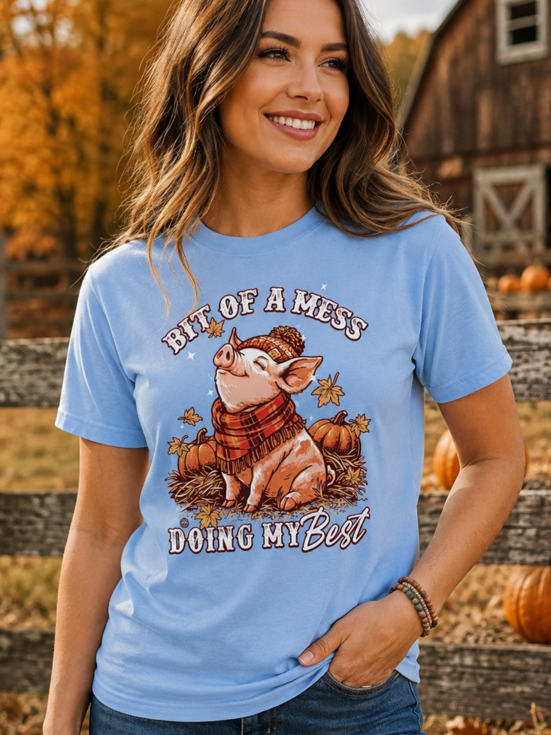 A woman wearing a light blue graphic t-shirt with a pig illustration and the text 'BIT OF A MESS DOING MY Best' in an autumn outdoor setting with a barn and pumpkins in the background.