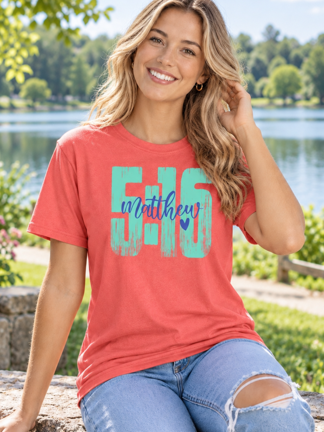 A woman sitting outdoors by a lake wearing a coral short-sleeve graphic T-shirt with large turquoise and navy text reading '5:16 Matthew' and light blue ripped jeans.