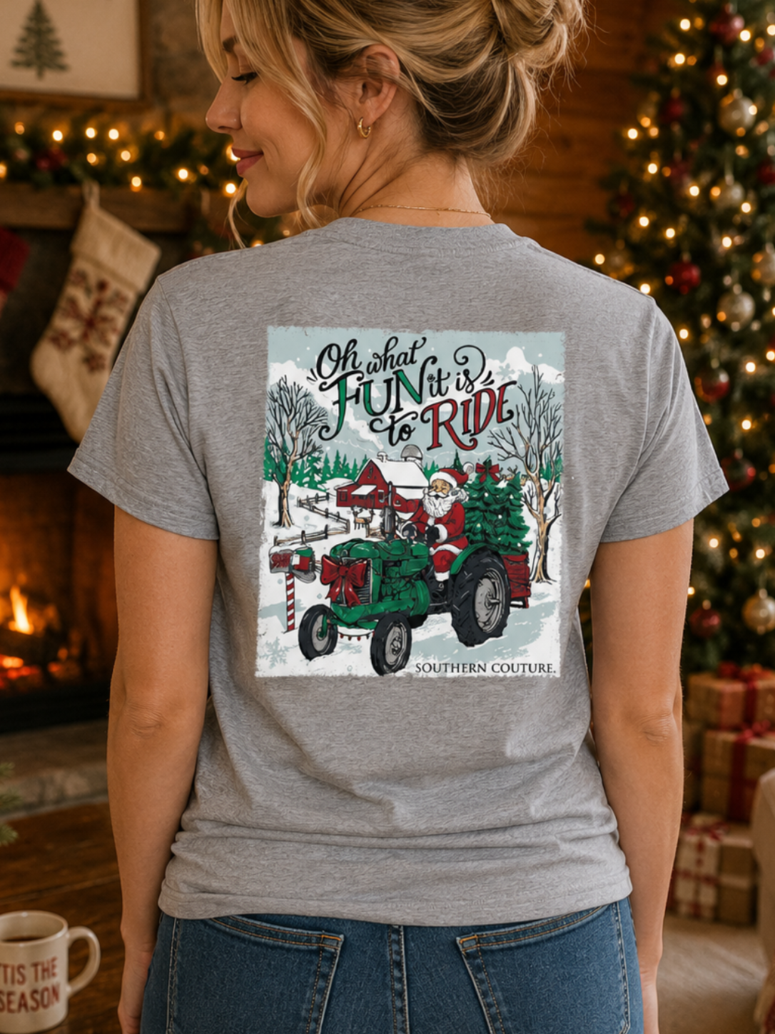 A woman wearing a heather gray graphic t-shirt with a Christmas tractor design, photographed from behind in a cozy, decorated living room with a fireplace and Christmas tree.