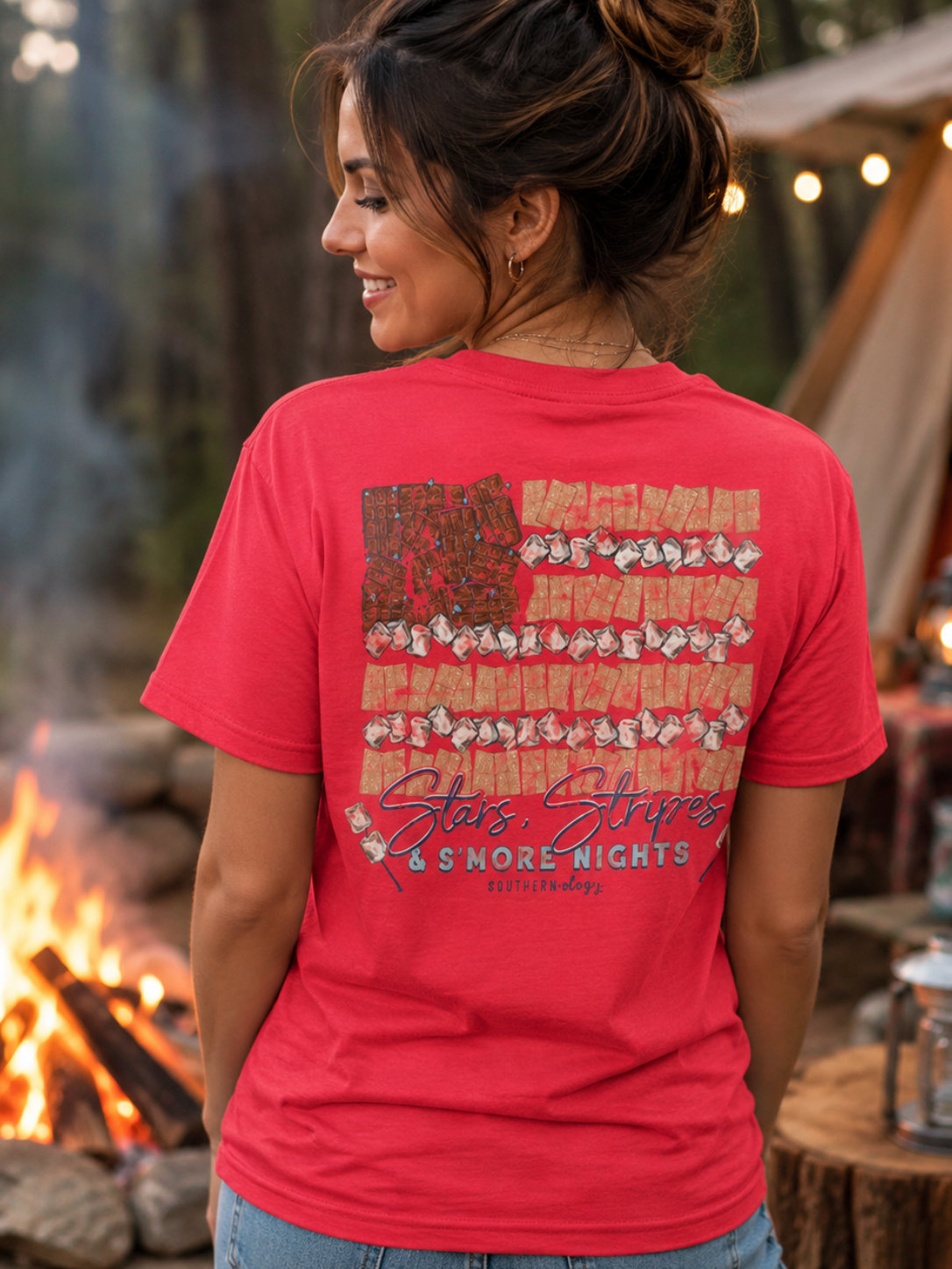 A woman wearing a red graphic t-shirt with a rustic American flag design featuring campfires and the text 'Star Stars &amp; MORE NIGHTS' on the back, photographed in an outdoor campfire setting.