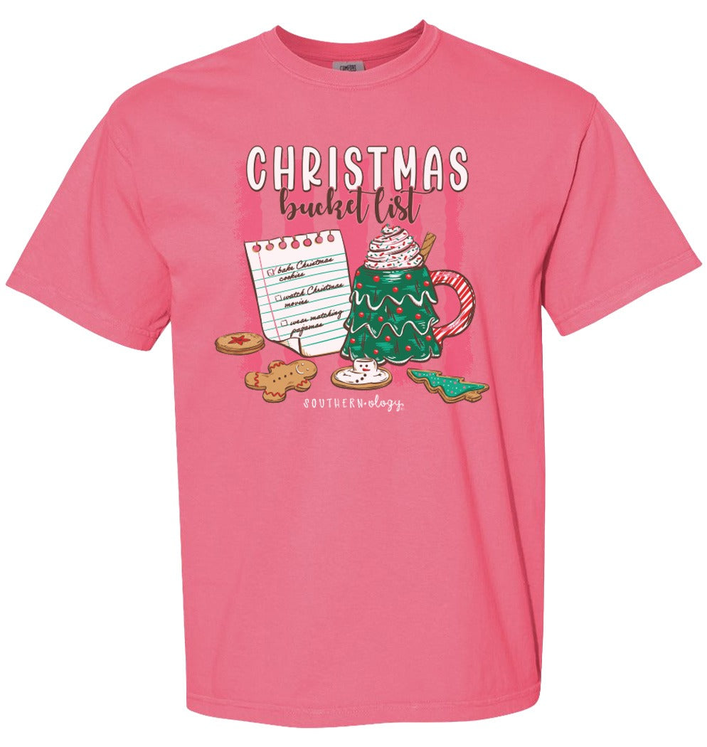 A pink short-sleeve crewneck t-shirt featuring a Christmas-themed graphic on the front with the text 'CHRISTMAS bucket list' above an illustration of a decorated Christmas tree, hot cocoa mug, cookies, and a notepad. The brand name 'southern dogg' is printed below the graphic.